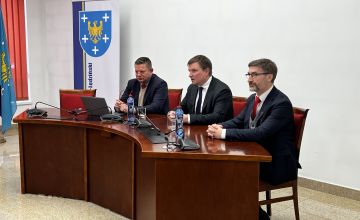 Trzy osoby siedzące przy półokrągłym stole. W rogu przy ścianie stoi ścianka z herbem powiatu.